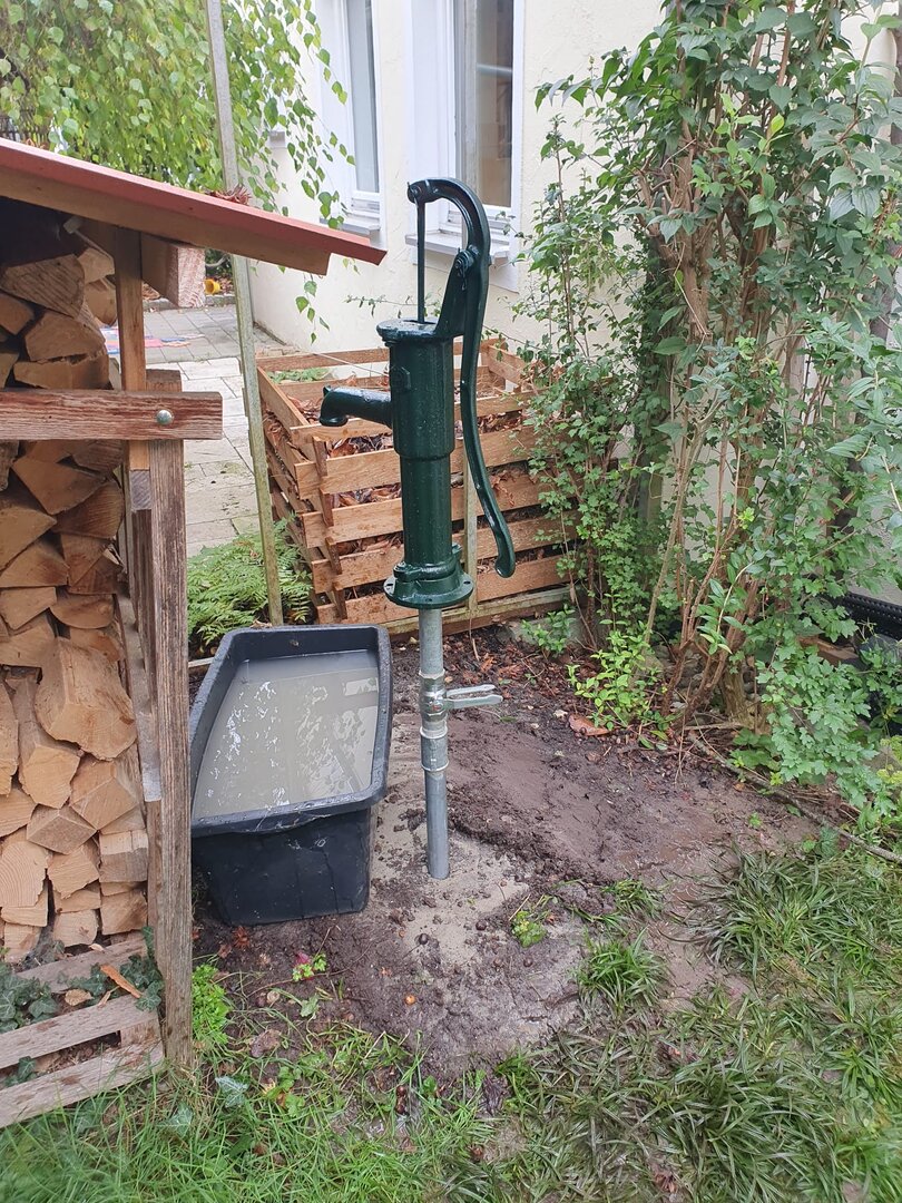 Schlagbrunnen mit Handpumpe in einem kleinen Gartenbereich